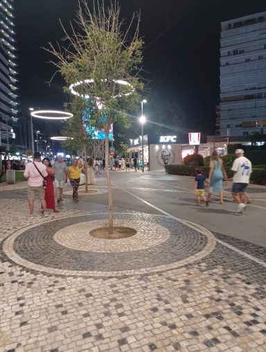 Pedestrians walk along a paved street at night, with a central tree and urban buildings.