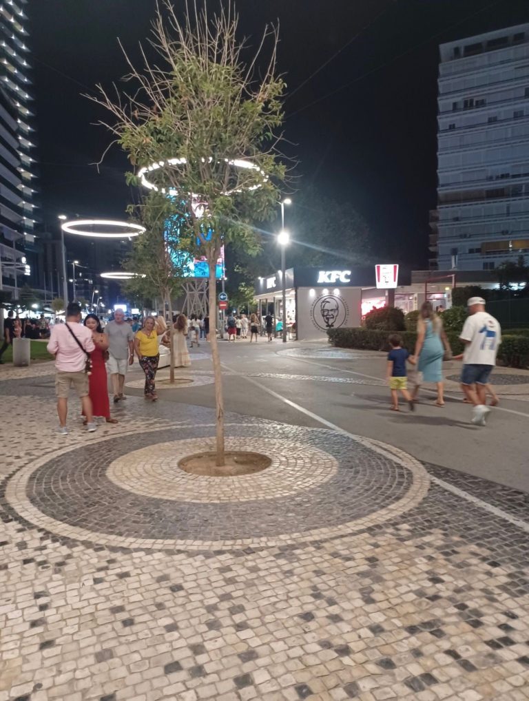Lively urban street at night with people walking and a central tree feature.